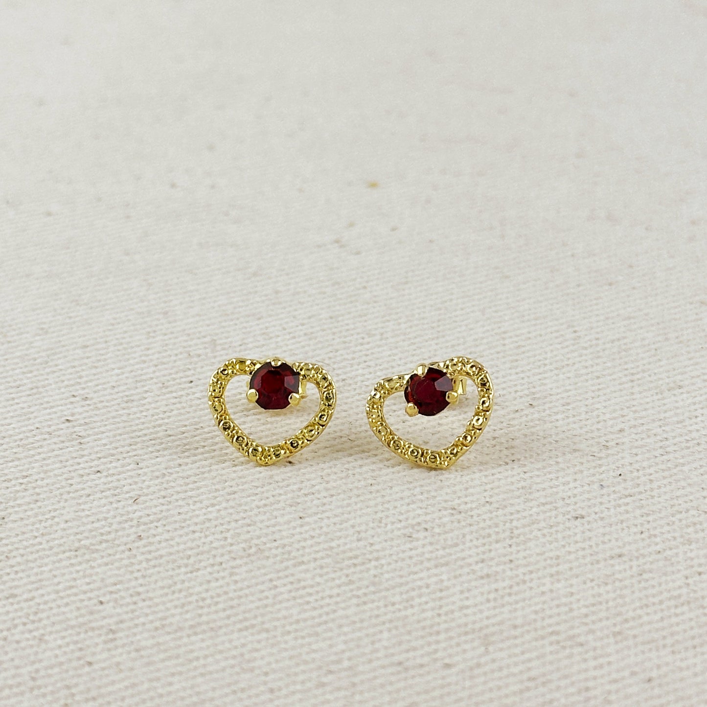 Gold heart-shaped earrings with red gemstones on a light beige background