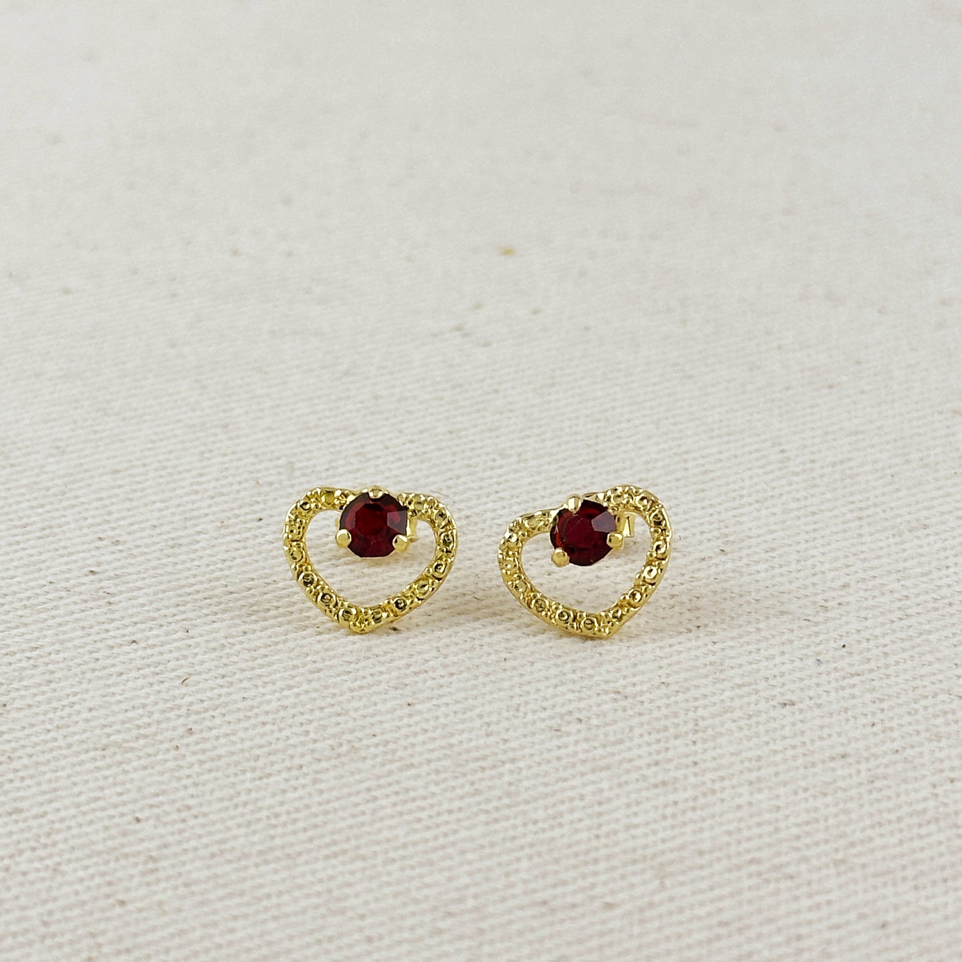 Gold heart-shaped earrings with red gemstones on a light beige background